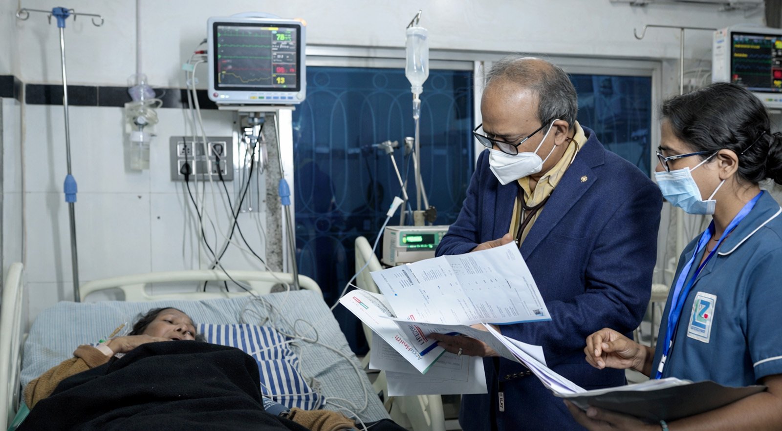 A doctor reviewing reports with a nurse beside a patient on a monitored bed in a nursing home.